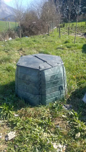 Contenedor hexagonal de plástico con tapa agrietada en pradera junto a árboles, imagen para arbizuko zabortegia