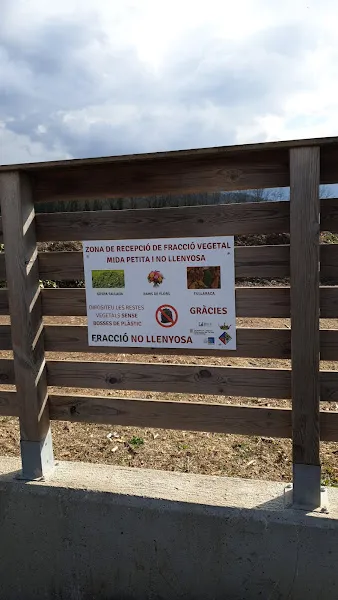 Recinto con cartel informativo de la area de compostatge comunitari de la fraccio vegetal junto a un vallado de madera y cielo nublado.