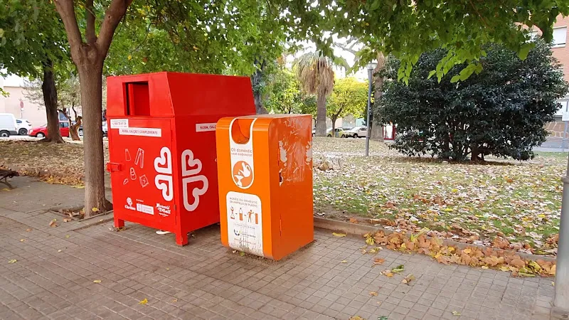 Contenedores rojo y naranja junto a un parque que señalan un punt de reciclatge d oli i roba para ropa y aceite doméstico