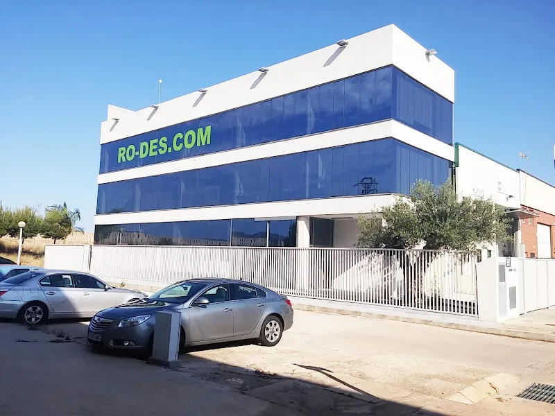Fachada de edificio de oficinas con cristales azules, coches aparcados y rótulo ro des en letras verdes.